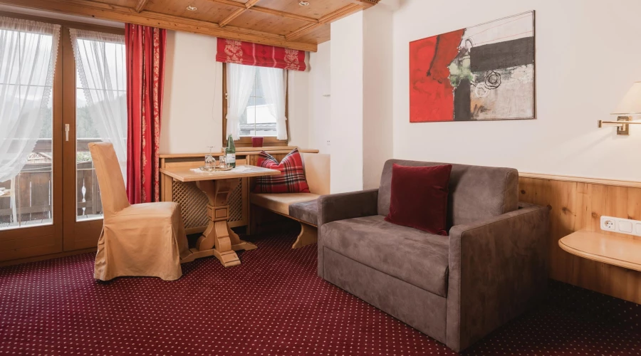 In der Junior Suite Edelweiss siehst du das Doppelbett und der kleinen Sitzecke und dem Sofa, im Hintergrund der balkonbereich.